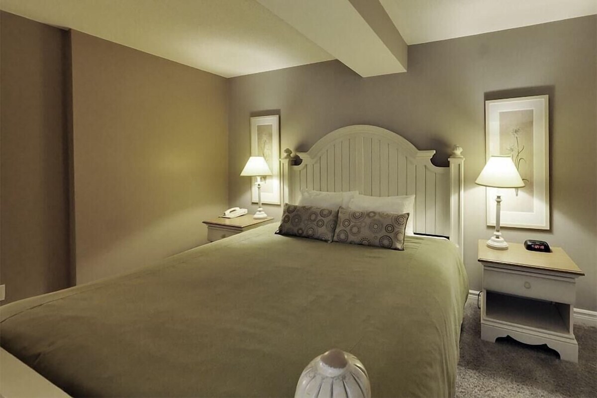 Ein ordentlich eingerichtetes Hotelzimmer mit einem großen Bett, Nachttischlampen, einem Nachttisch und einem gerahmten Bild an der Wand.