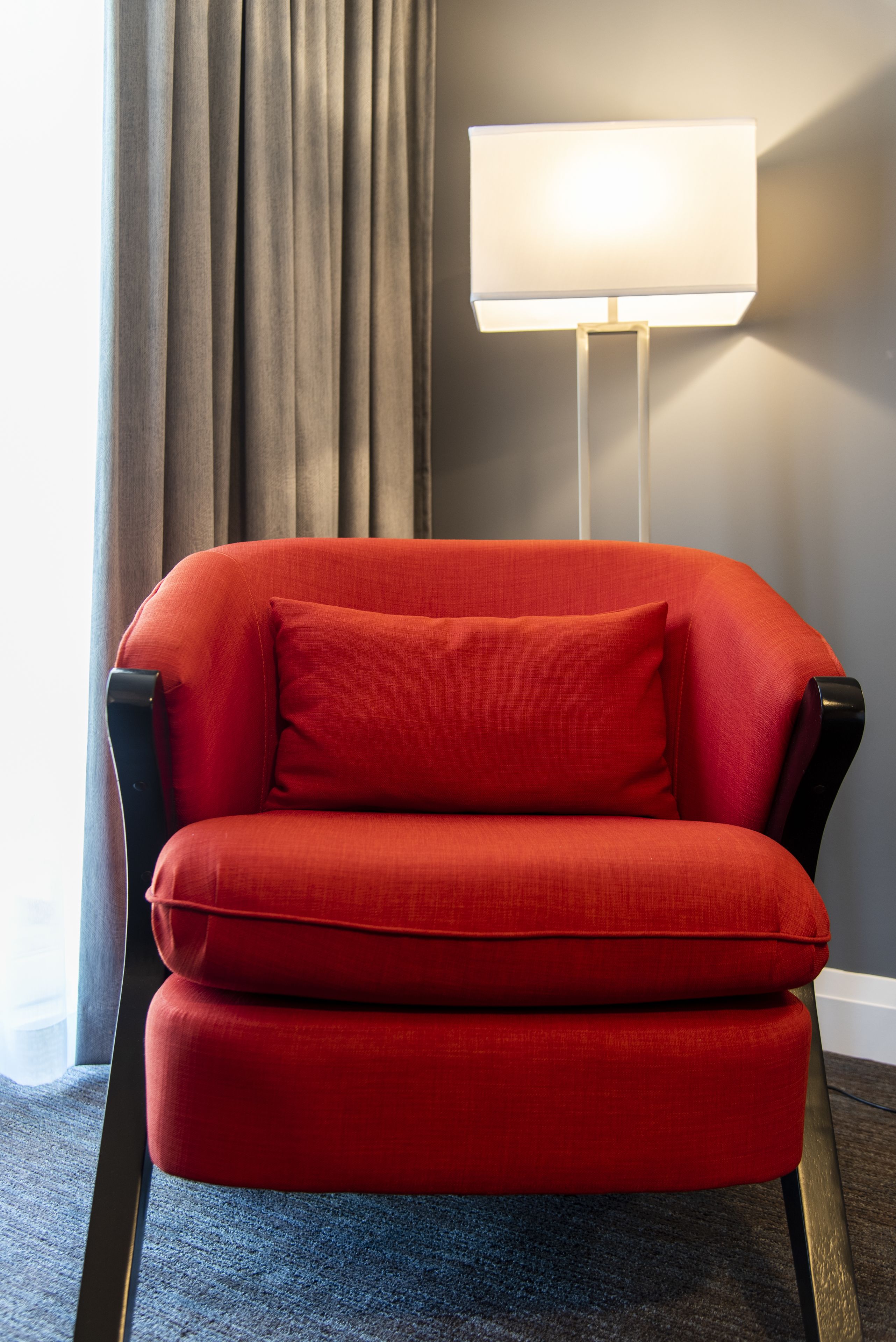 A red armchair with a matching pillow in a room with a grey curtain and a white floor lamp.
