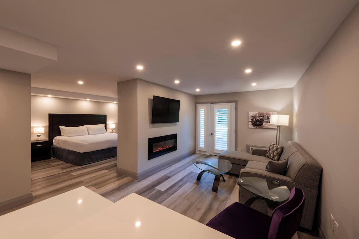 Een moderne hotelkamer met een bed, een aan de muur bevestigde televisie, een open haard, een glazen tafel en een zithoek met een bank en fauteuil.