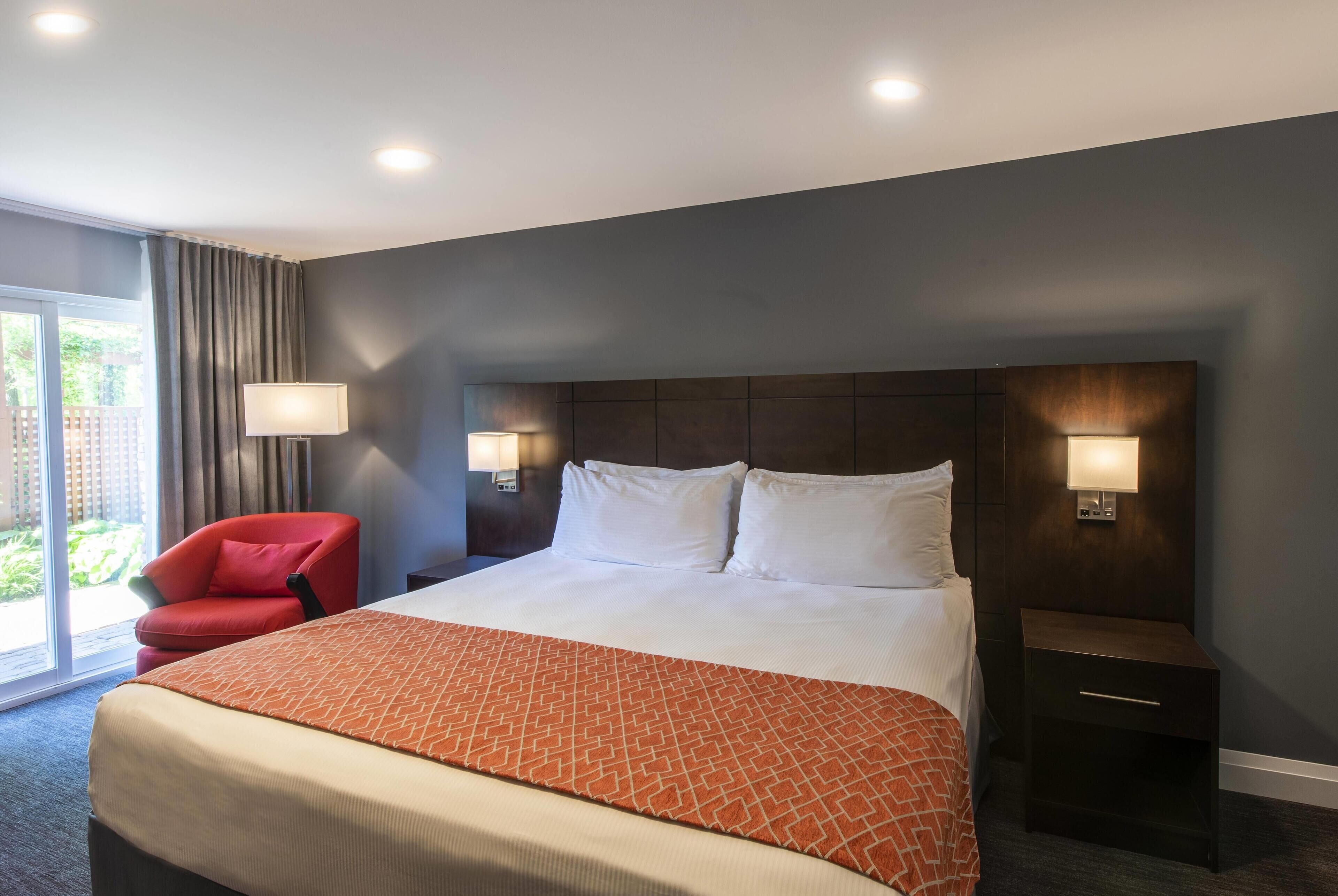 A hotel room with a large bed, two bedside lamps, a red chair, and a view of the outdoors.