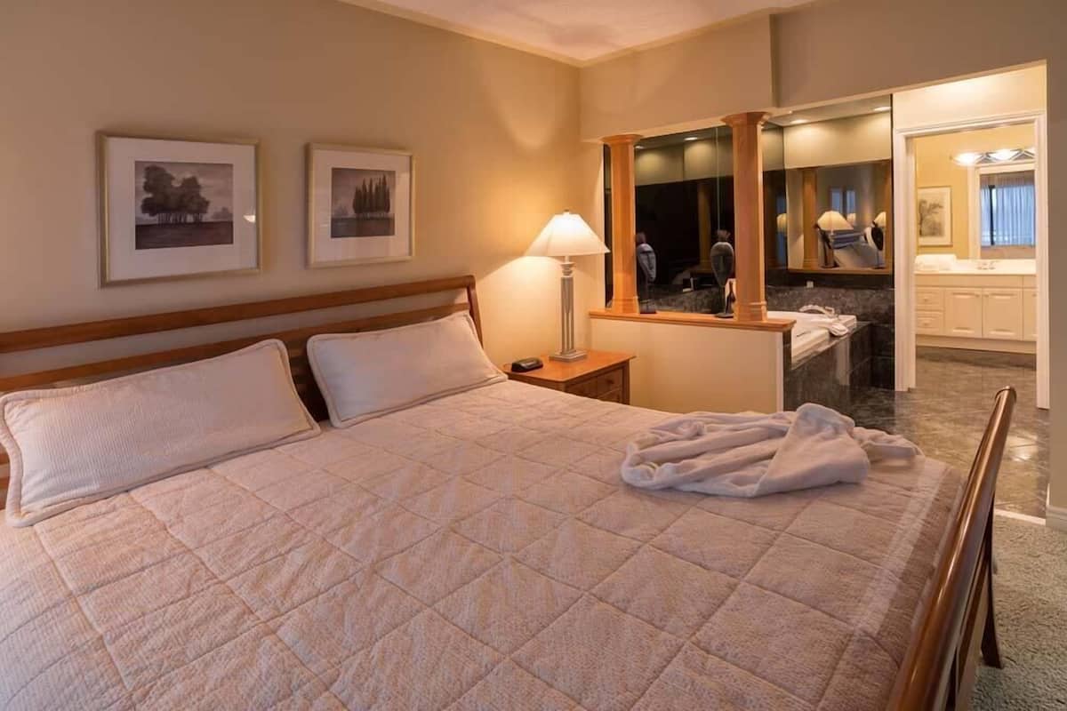 Een hotelkamer met een groot bed, een nachtkastje met een lamp en een spiegel. Er hangen foto's in lijsten aan de muur en er is een badkamer zichtbaar door een geopende deur.
