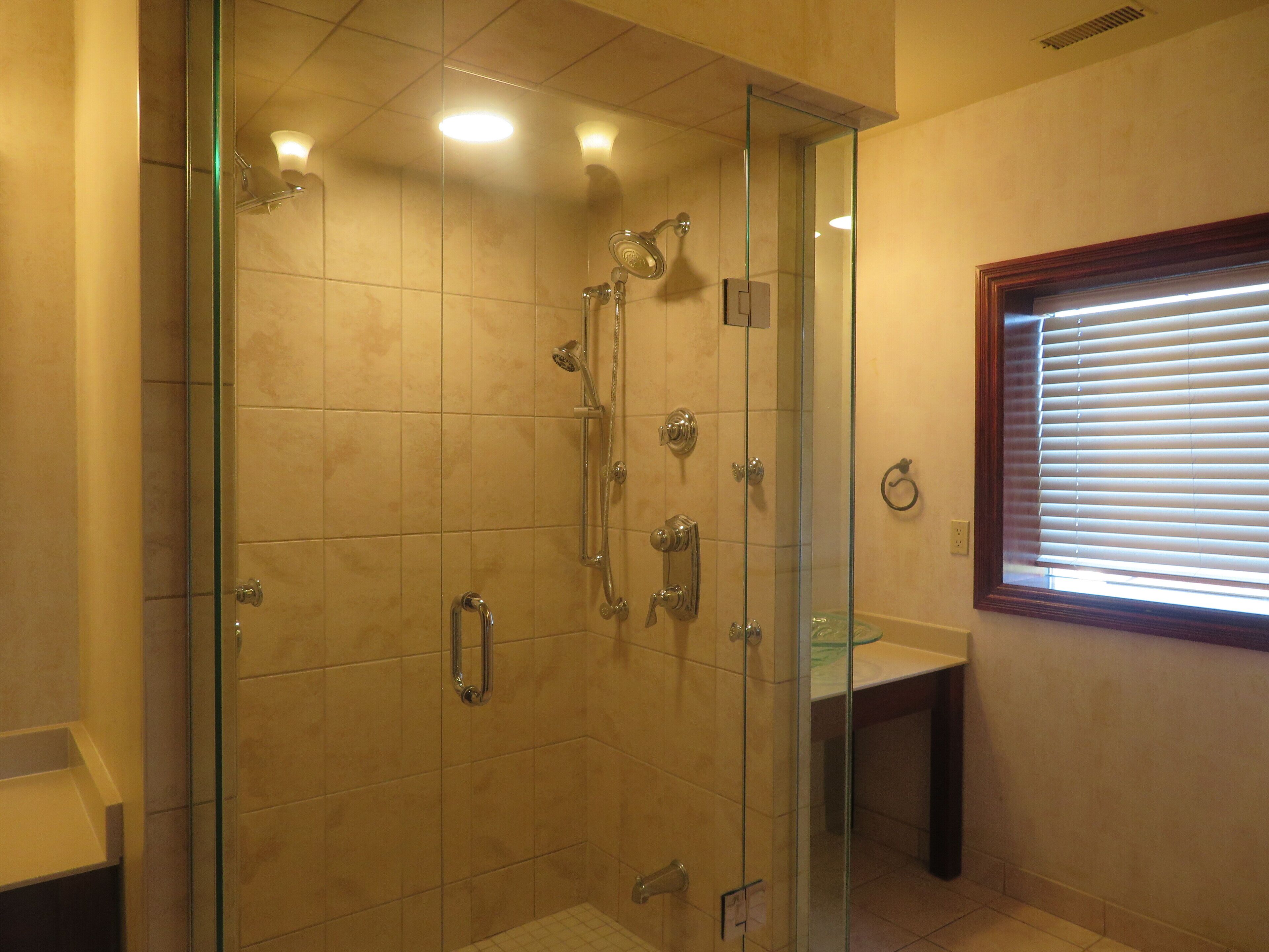 A glass-enclosed shower with multiple fixtures, a bench, and a window with blinds.