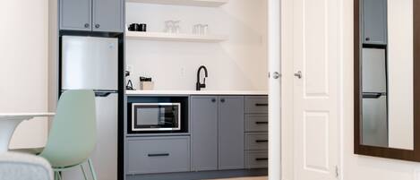 Cocina compacta con armarios grises, microondas, pileta y un pequeño comedor con una silla.