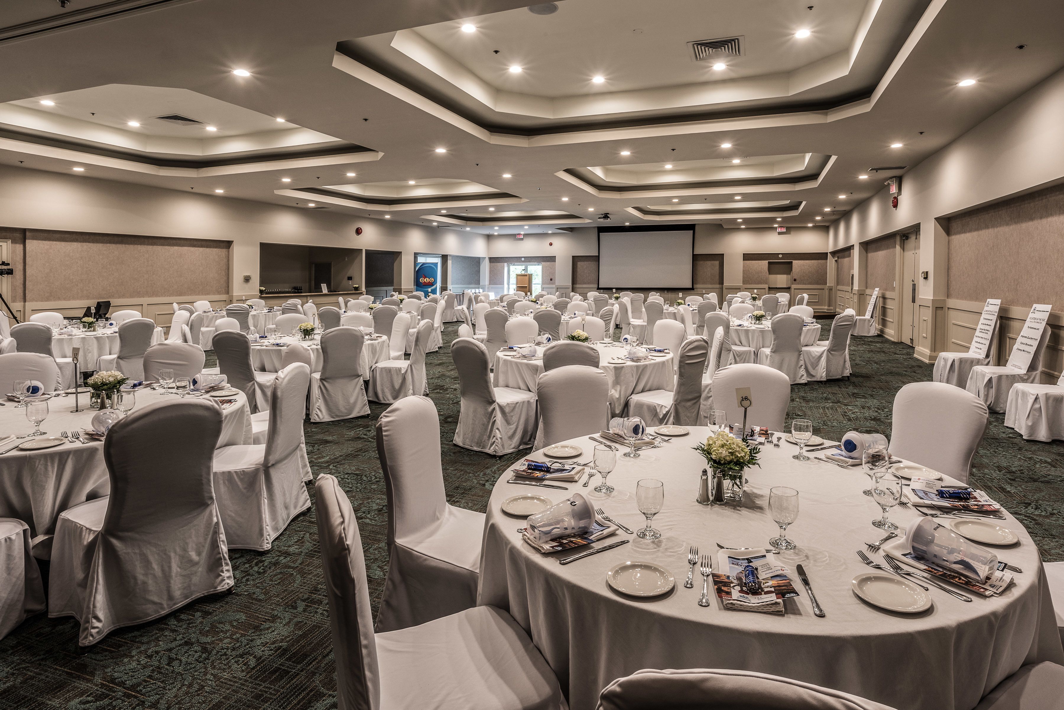 A large banquet hall with round tables set for dining, white chairs, and a carpeted floor.