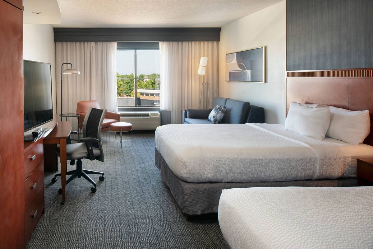 A hotel room with a large bed, a desk with a TV, a chair, and a window with curtains.