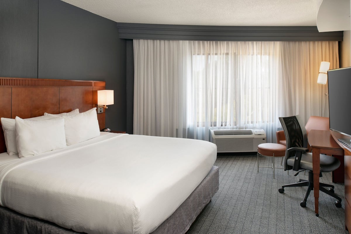 A hotel room with a large bed, a desk with a chair, an air conditioning unit, and a television.