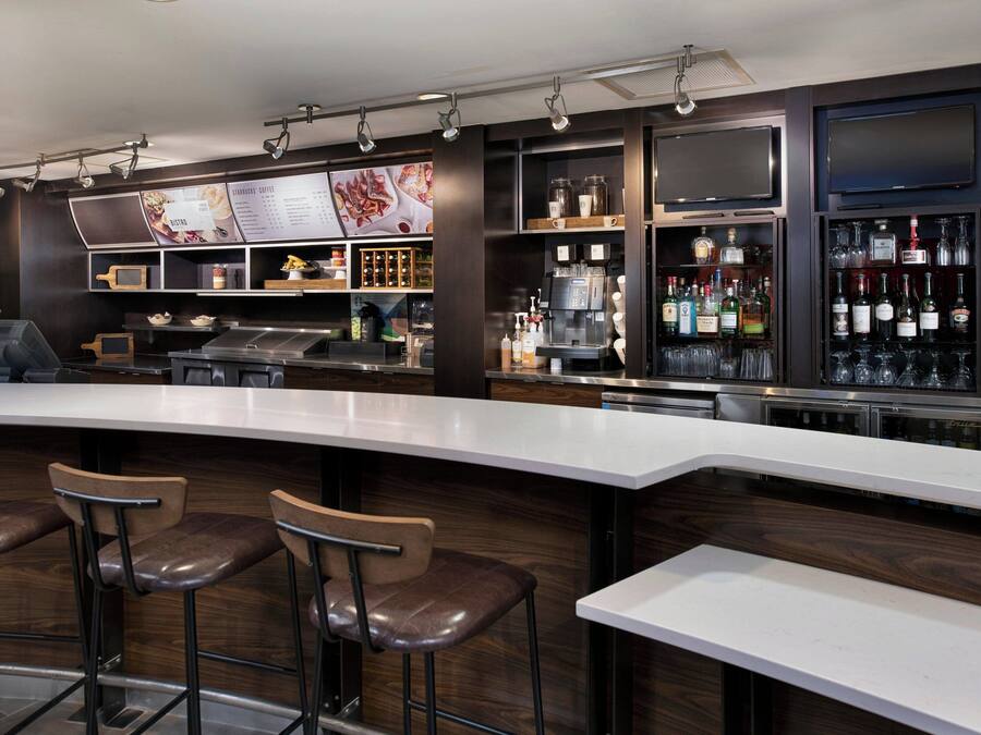 Een moderne bar met een gebogen toonbank, barstoelen, een koffieapparaat en diverse drankflessen.