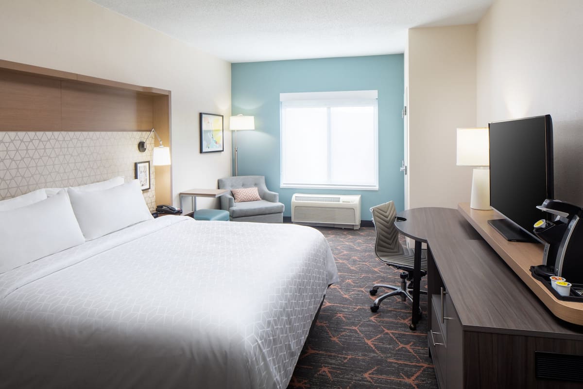 A hotel room with a large bed, a desk with a computer, a chair, a TV, and a window with blinds.