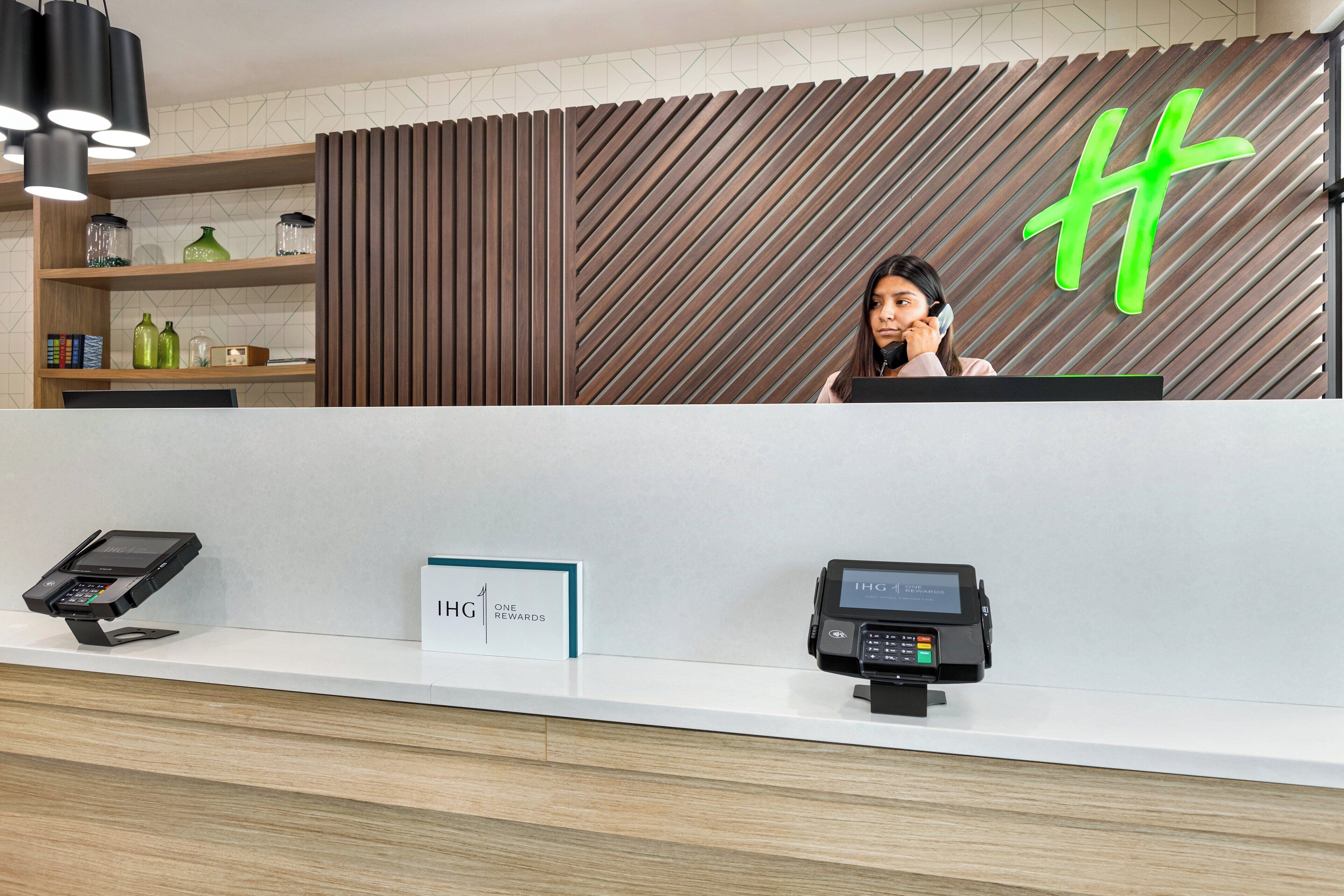 Zona de recepción de un hotel con diseño moderno, que incluye un mostrador de madera, un terminal de pago digital y un cartel que anuncia un programa de recompensas.