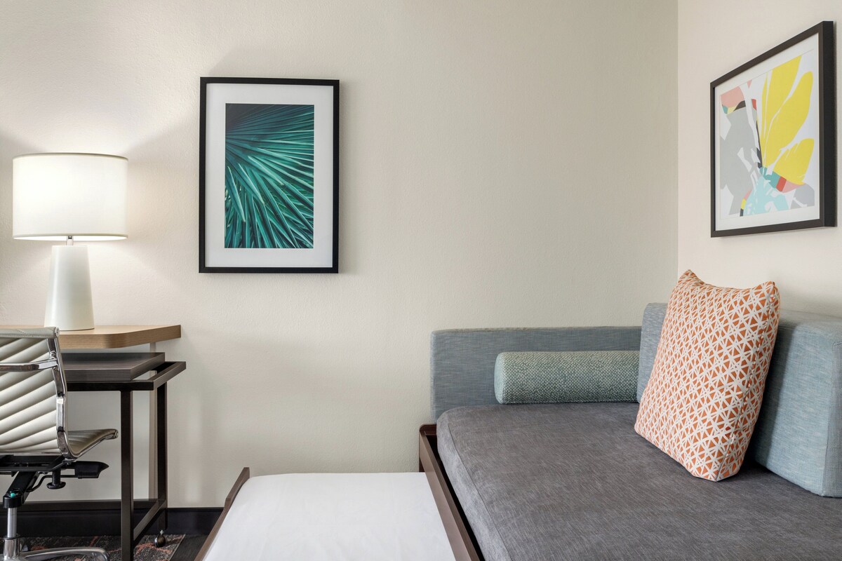A hotel room with a bed, a desk with a lamp, and two framed artworks on the wall.