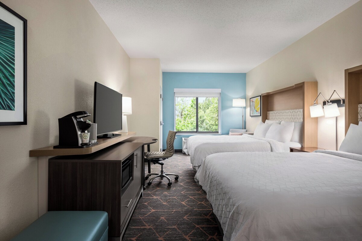 A hotel room with two beds, a desk with a TV, a chair, and a window with a view of trees.