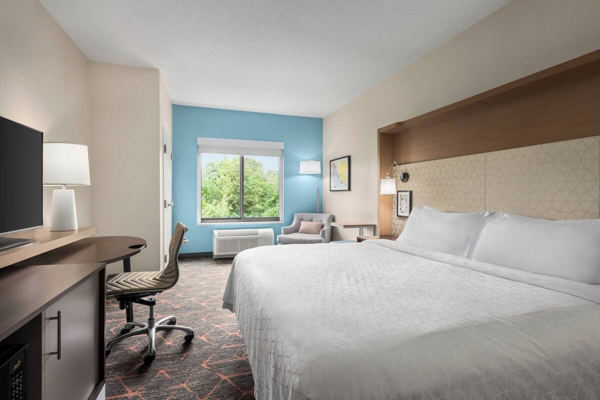 A hotel room with a bed, a desk with a lamp, a chair, and a window with a view of trees.