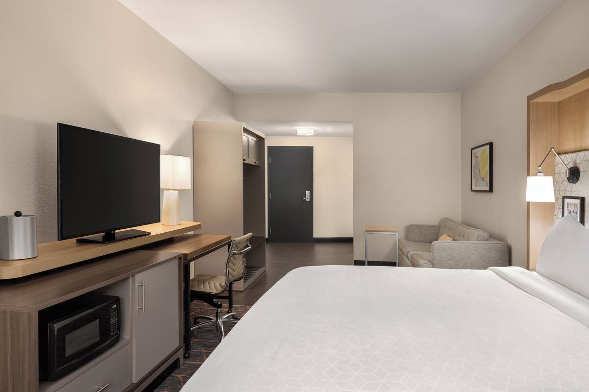 A hotel room with a bed, a TV, a desk with a chair, a microwave, a door, and a small table with a lamp.