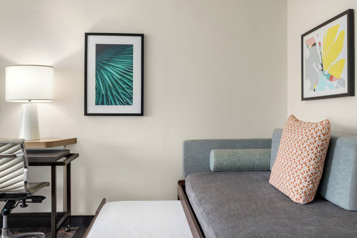 A hotel room with a bed, a desk with a lamp, and two framed artworks on the wall.