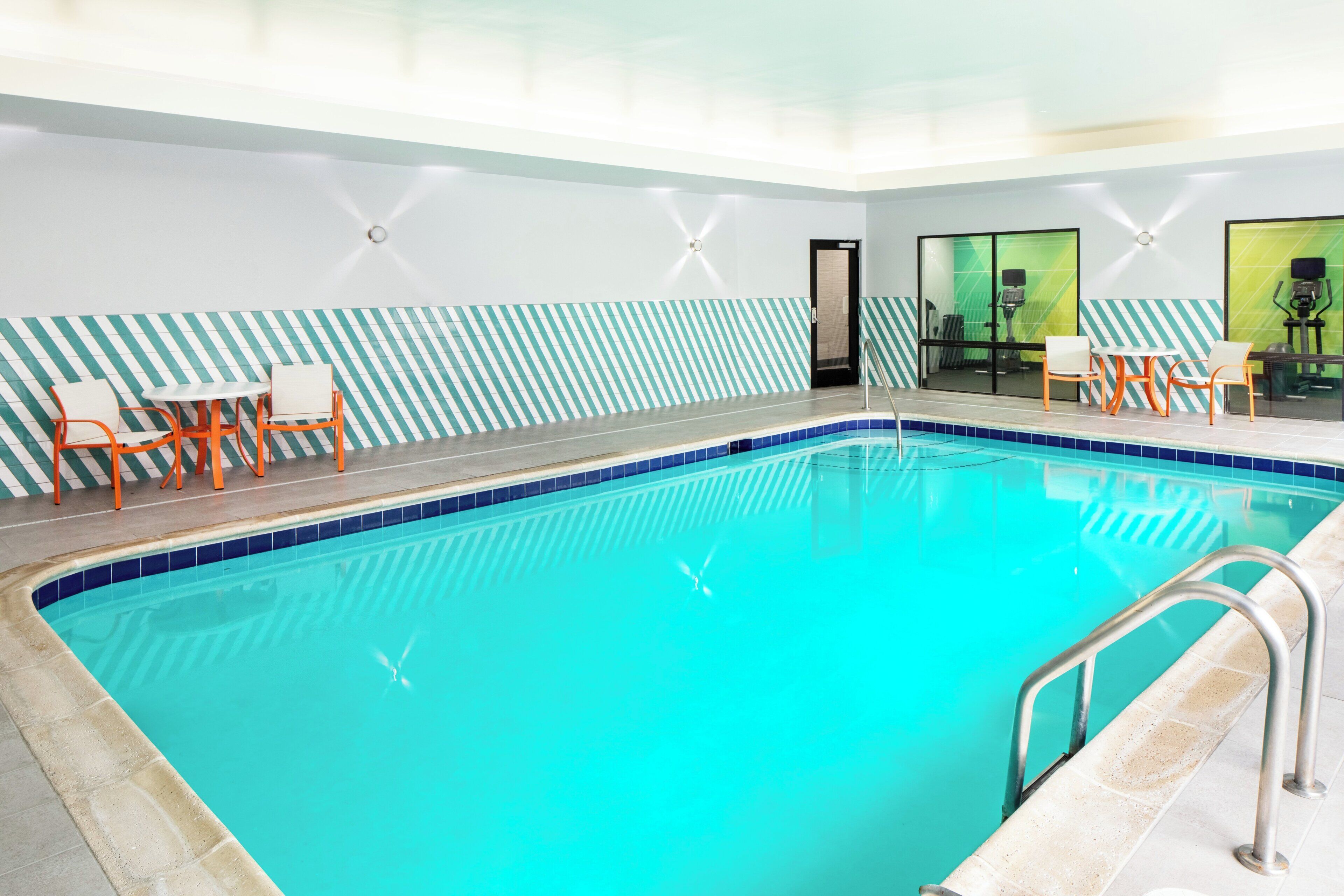 An indoor swimming pool with a tiled edge, surrounded by orange chairs and tables, and a striped wall pattern.