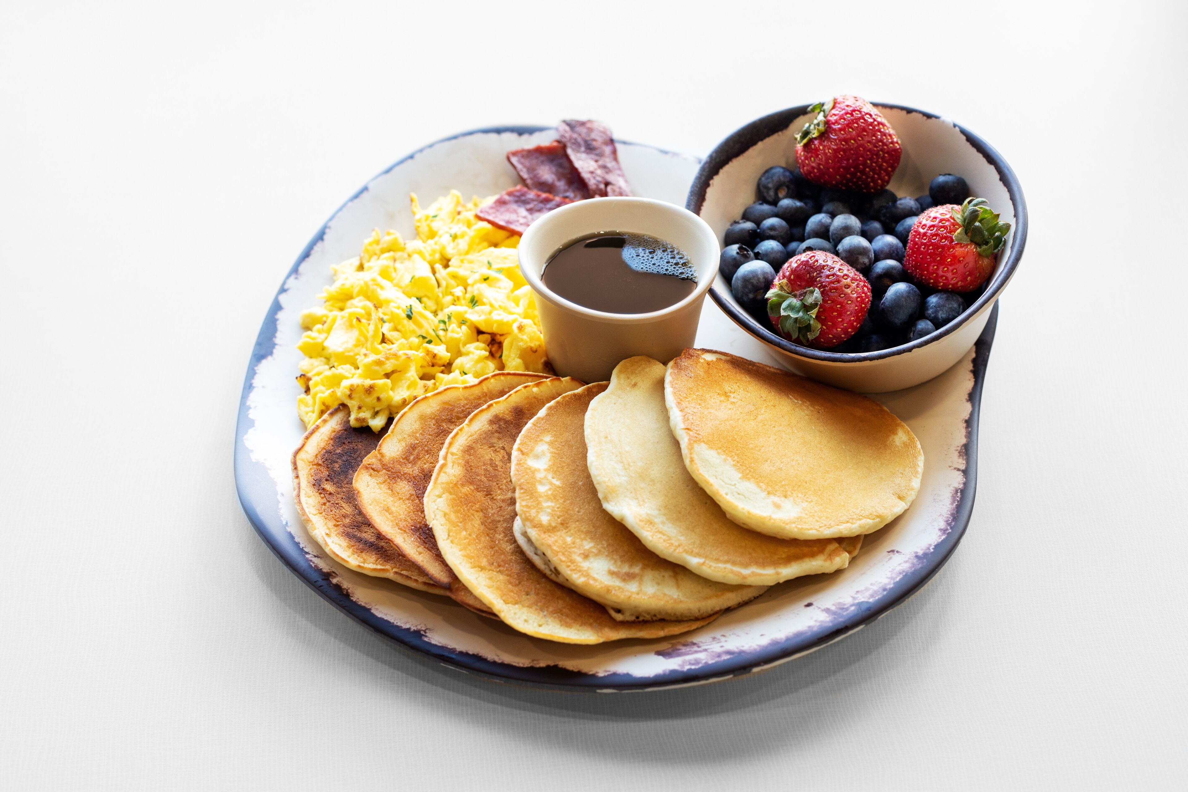 A breakfast plate with pancakes, scrambled eggs, bacon, blueberries, strawberries, and a cup of coffee.