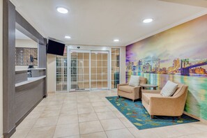 Eine Hotelhalle mit einer Rezeption, einem Flachbildfernseher, einer Sitzecke mit zwei Sofas, einem gemusterten Teppich und einem Wandgemälde einer Stadtansicht.