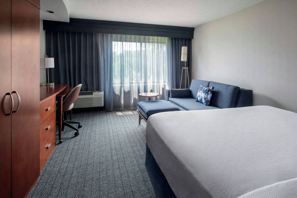 A hotel room with a large bed, a desk with drawers, a chair, a small round table, and a floor lamp.