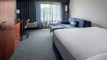 A hotel room with a large bed, a desk with drawers, a chair, a small round table, and a floor lamp.
