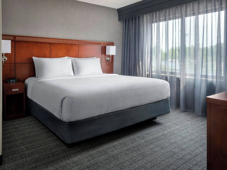 A hotel room with a large bed, two bedside tables with lamps, a wooden headboard, and a view of the outdoors through sheer curtains.