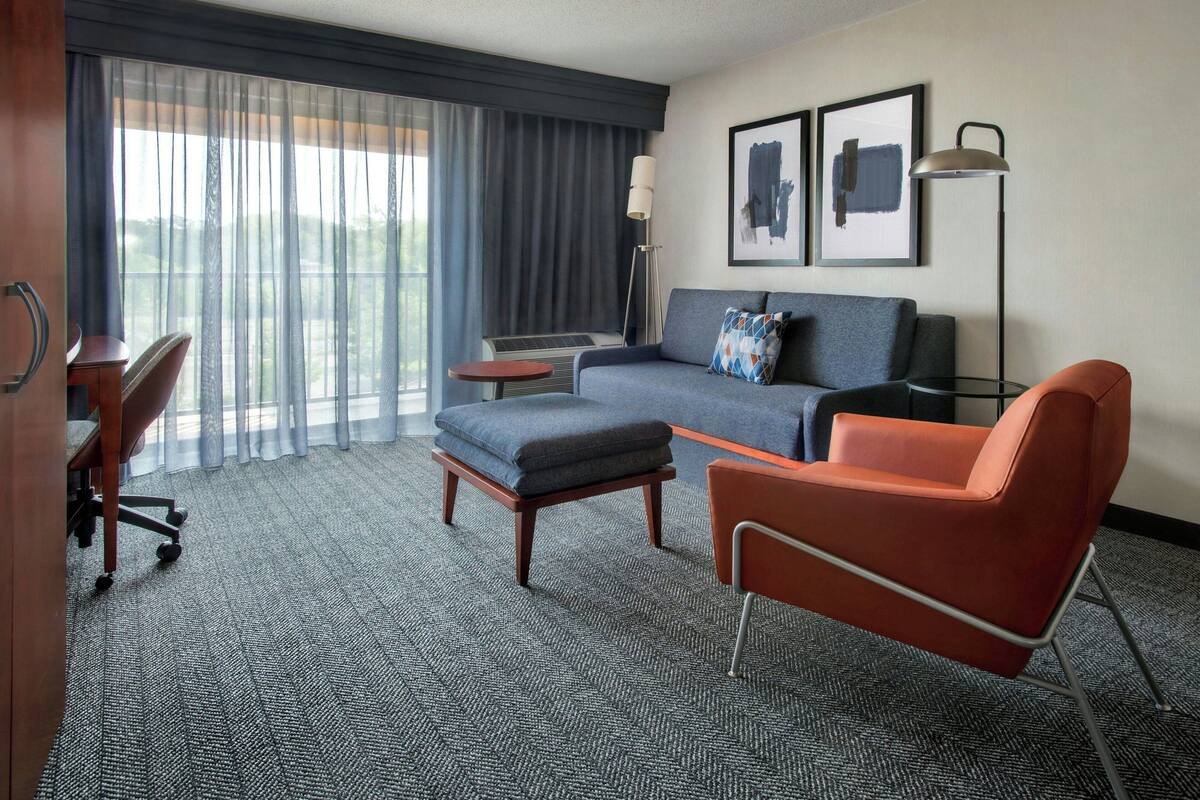 A hotel room with a sofa, a red chair, a small table, a desk with a chair, and a large window with curtains.