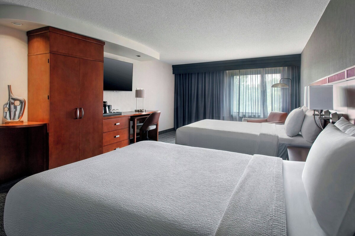 A hotel room with two beds, a desk with a chair, a TV, and a large window with curtains.
