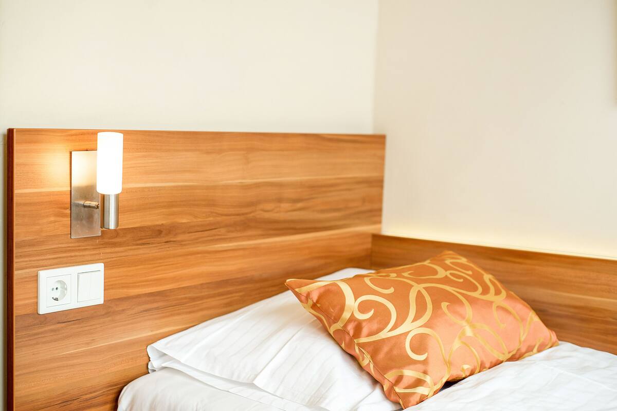 Een net opgemaakt bed met een witte matras en een decoratief kussen, een wandlamp en een stopcontact op een houten hoofdbord.
