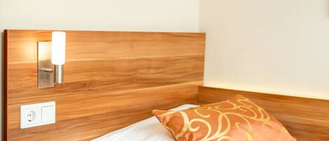 A neatly made bed with a white mattress and a decorative pillow, a wall-mounted light fixture, and a power outlet on a wooden headboard.