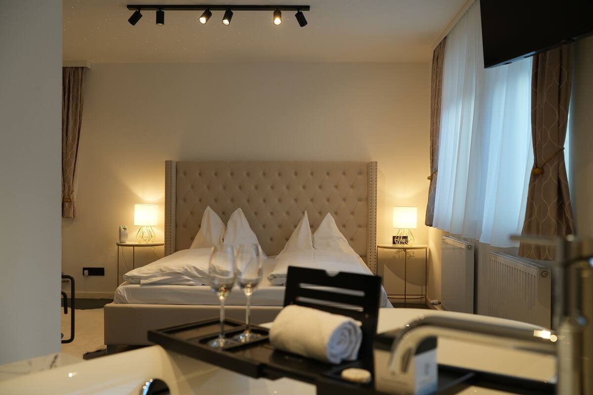 Hotelkamer met een groot bed, twee bedlampen, een dienblad met glazen en een handdoek, en een raam met gordijnen.