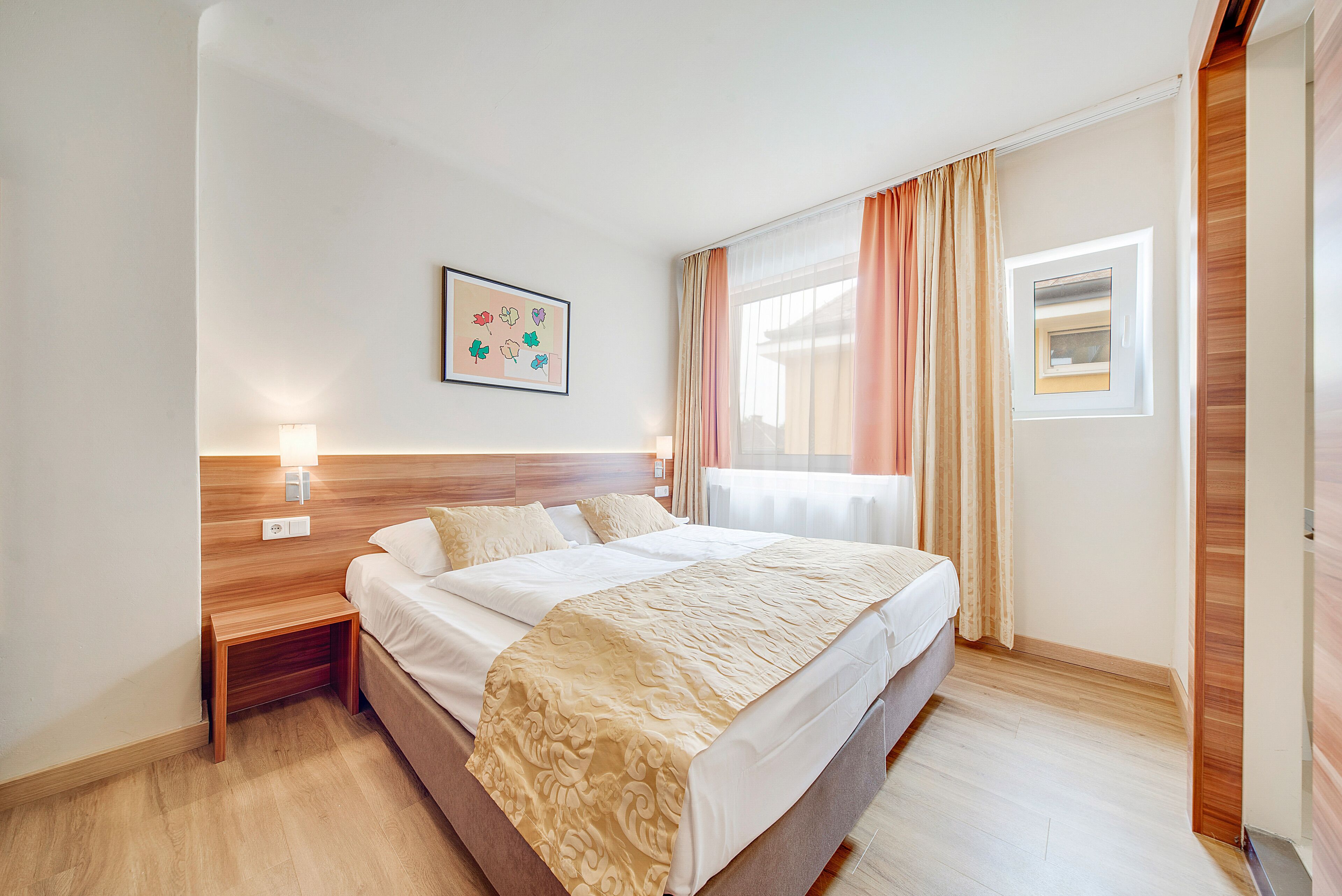 A hotel room with a bed, bedside tables, a wall-mounted light, a window with curtains, and a small skylight.