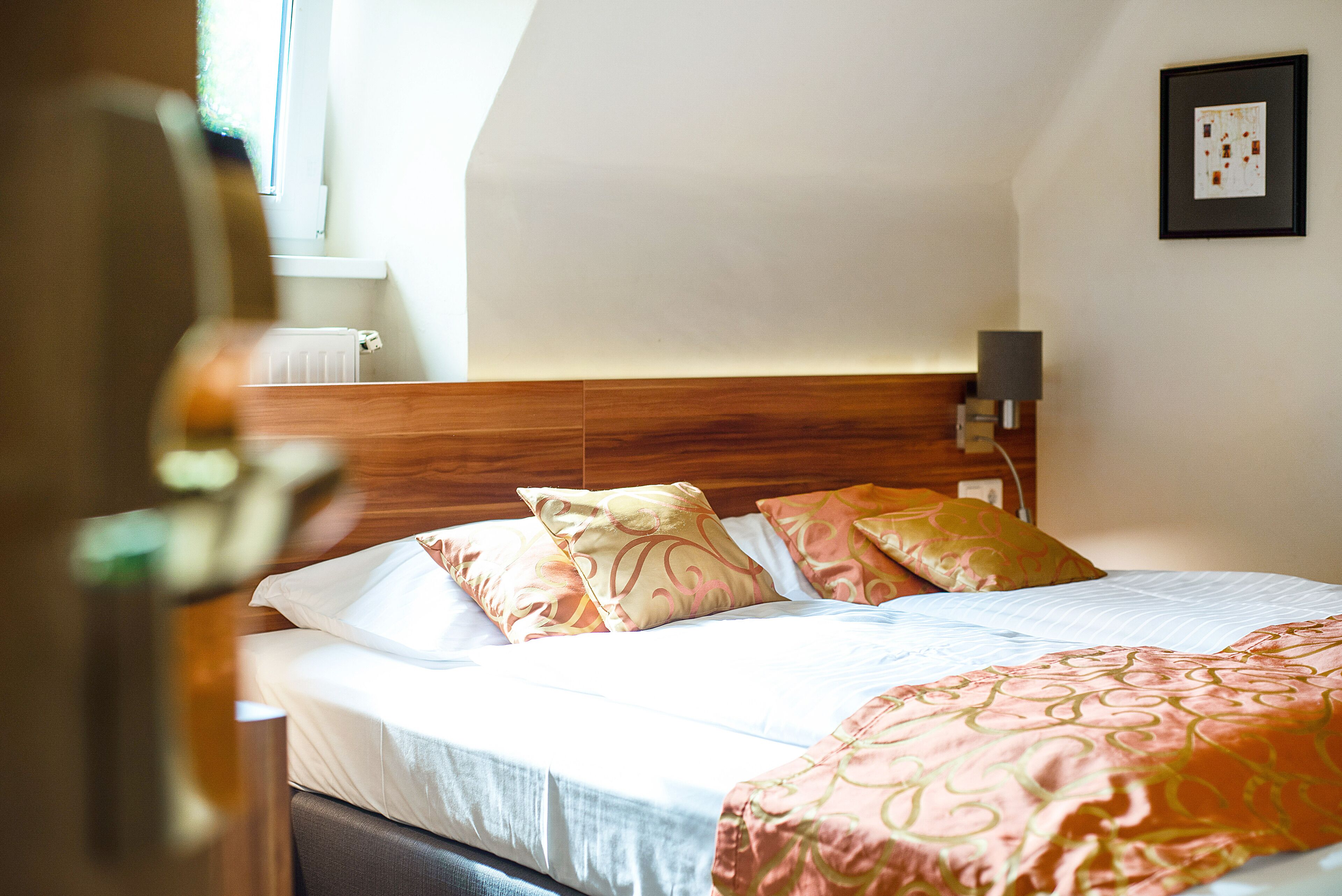 Una cama bien hecha con almohadillas estampadas y un cabecero, una lámpara de noche y un cuadro en la pared.