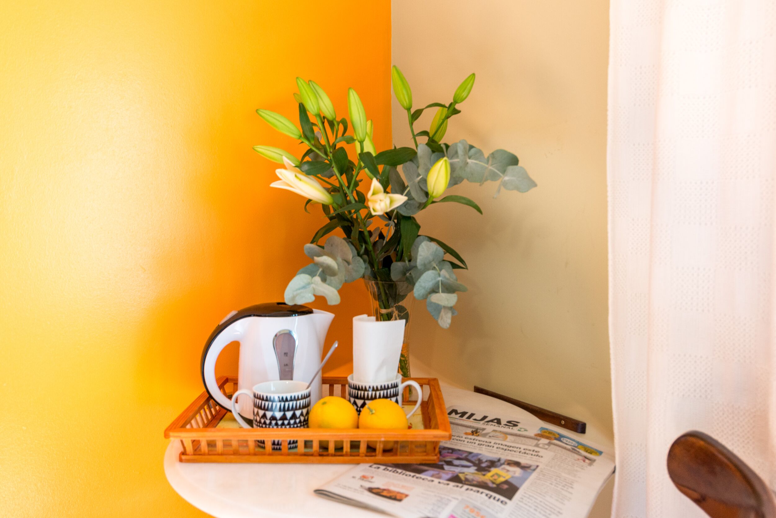 A corner of a room with a table holding a tray with a teapot, cups, and lemons, a vase with flowers, and a newspaper.