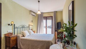 A hotel room with a bed, bedside table, lamp, air conditioning unit, curtains, a television, a plant, and a table with a wine glass and book. - Hostel El Escudo de Mijas (Mijas)