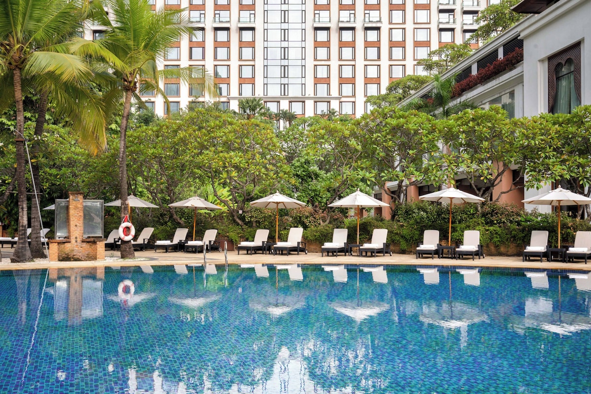 Piscina com espreguiçadeiras e guarda-sóis, rodeada por vegetação exuberante e um edifício alto ao fundo.