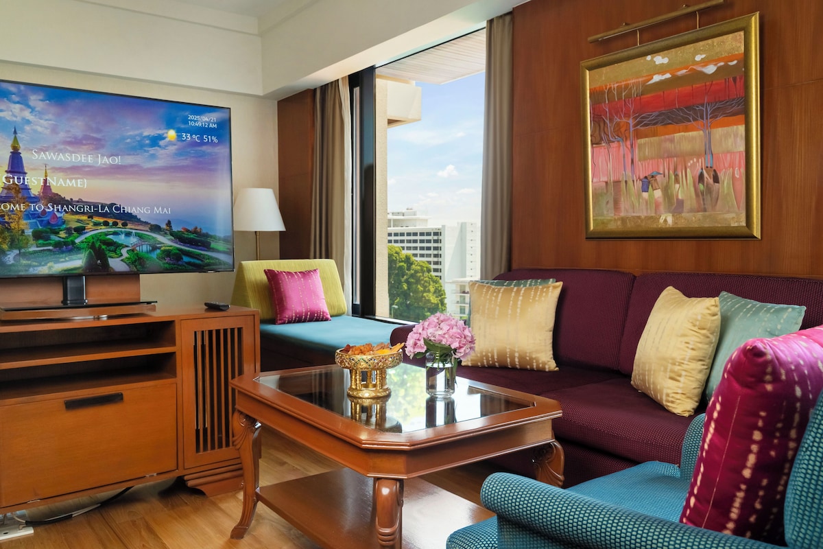 Quarto de hotel com televisão exibindo uma vista panorâmica, sofá com almofadas coloridas, mesa de centro com uma tigela decorativa e um quadro na parede.