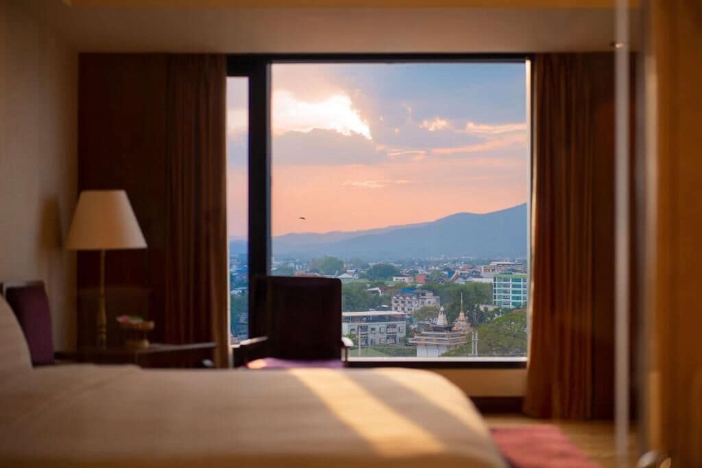 A hotel room with a large window, a bed, a chair, and a lamp.