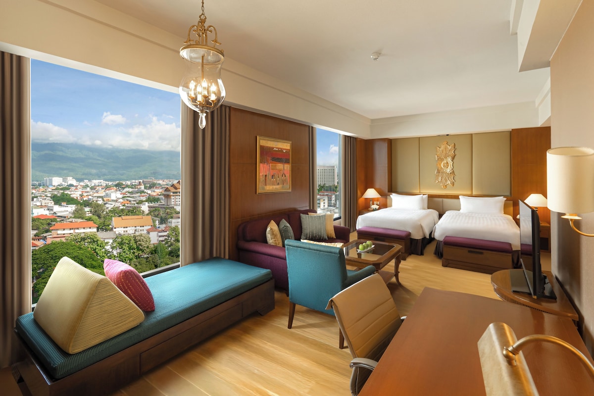 Quarto de hotel com vista para a cidade, um sofá, duas camas, uma escrivaninha e um lustre.