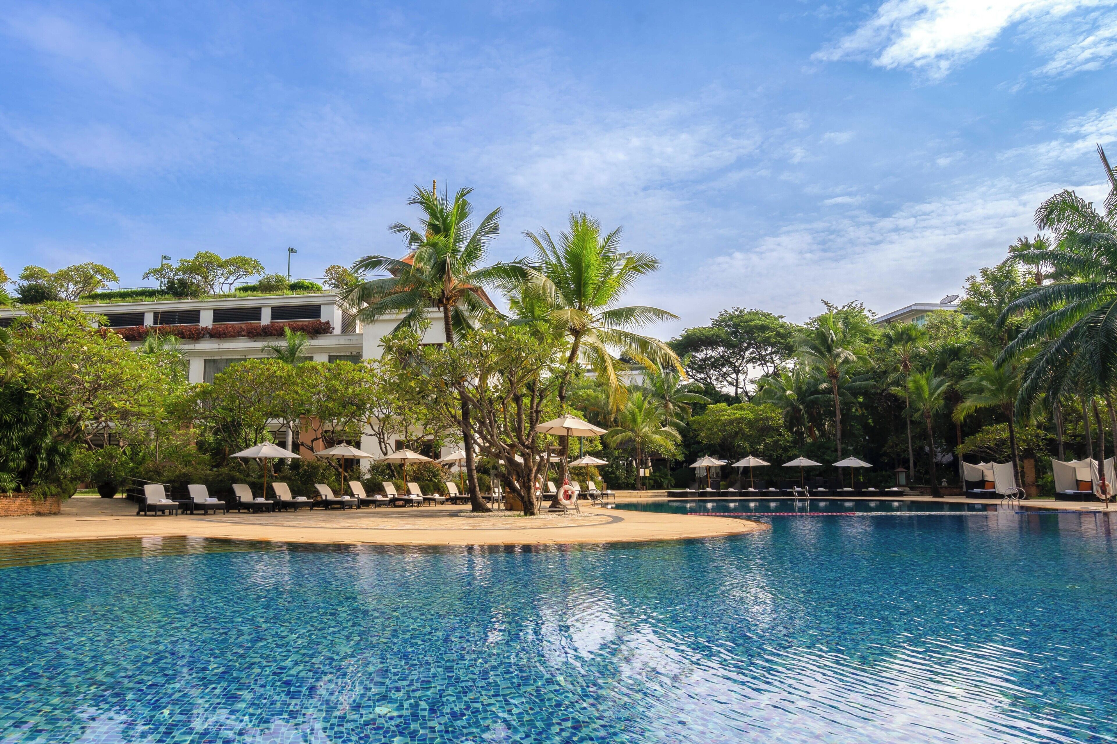 A clear blue pool with lounge chairs and umbrellas, surrounded by palm trees and a building with a flag.