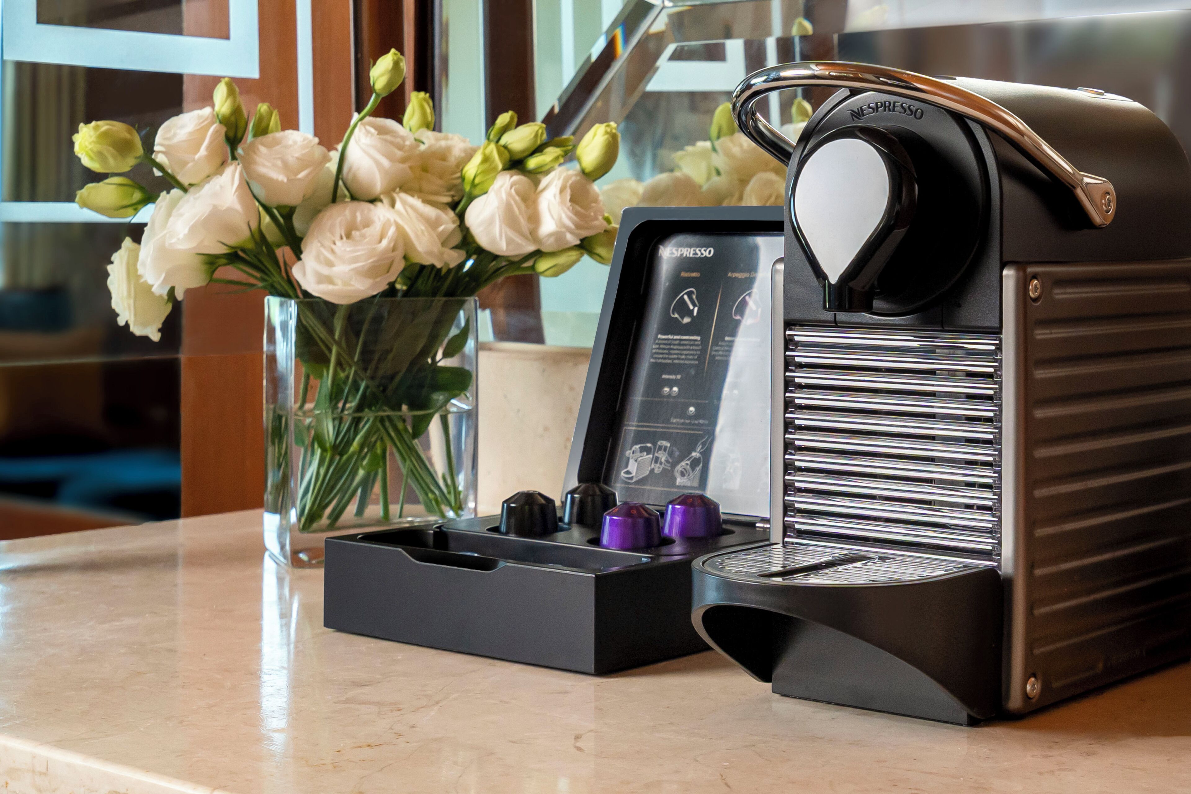 Una máquina de café Nespresso con un portacapsulas y un jarrón con flores blancas sobre una encimera de mármol.