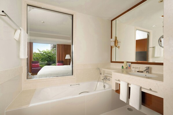 A modern bathroom with a large bathtub, a vanity with a mirror, and a window offering a view of a bed and a chair outside.