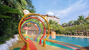 A swimming pool with a colorful play structure in the foreground and a hotel in the background.
