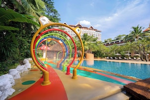 Una piscina con una estructura de juego colorida en primer plano y un hotel al fondo.