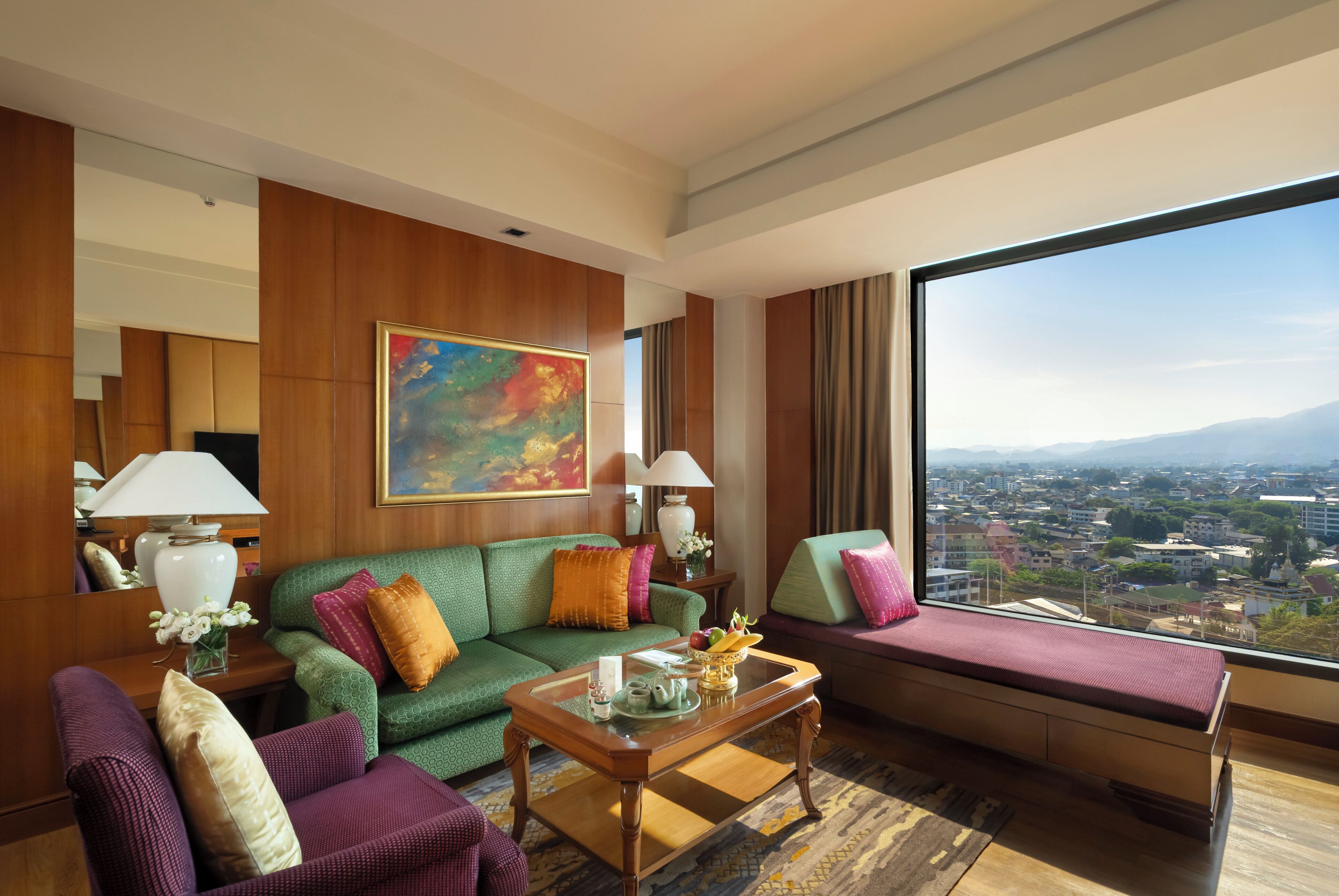 A hotel room with a green sofa, a purple armchair, a wooden coffee table, a painting on the wall, and a large window with a city view.
