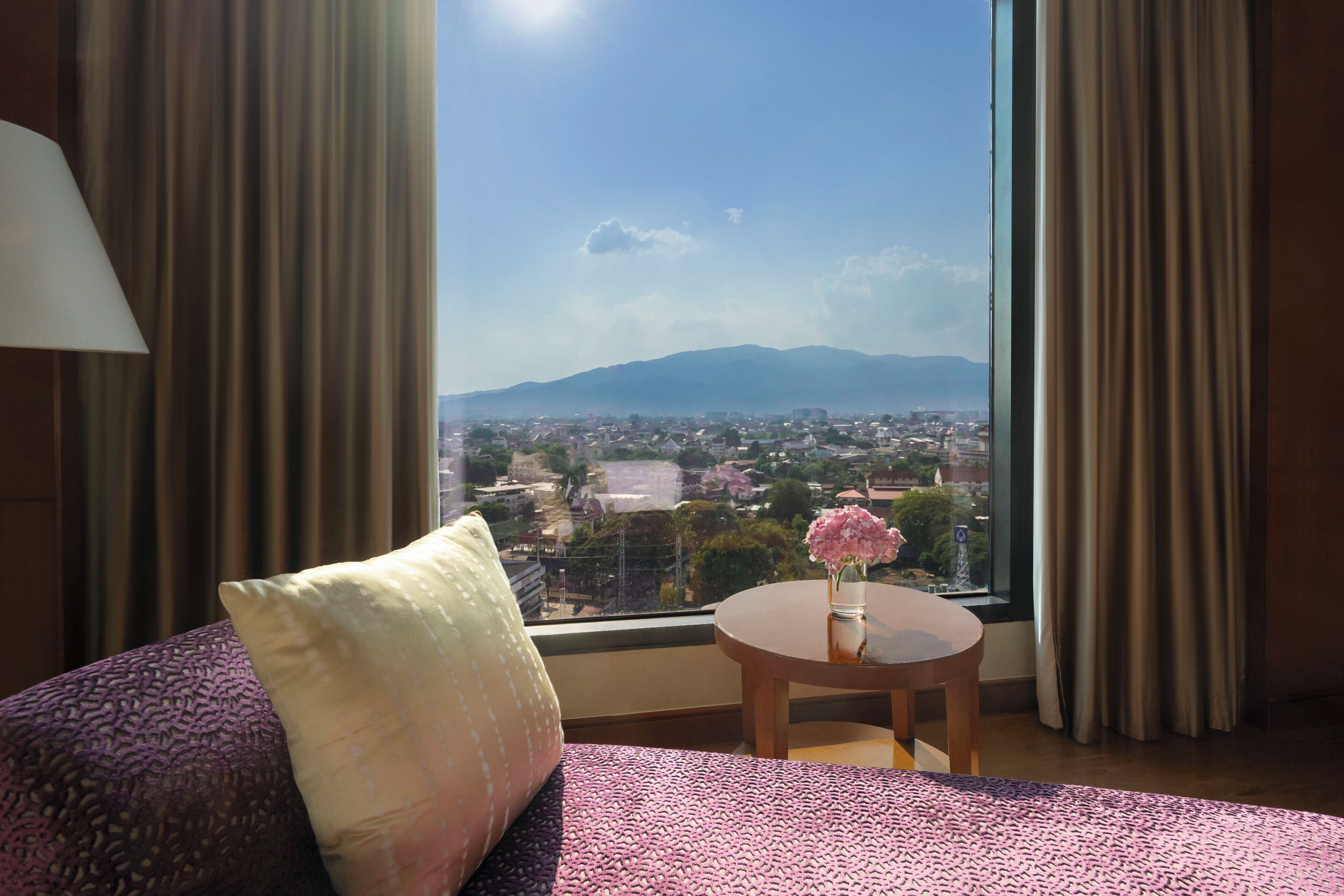 A hotel room with a bed, a small table with a vase of flowers, and a view of a cityscape and mountains through a large window.
