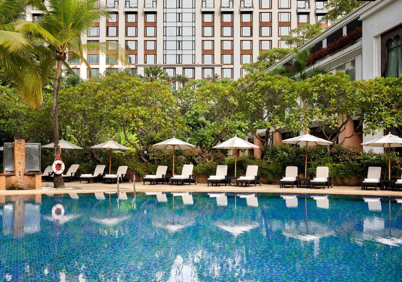 A swimming pool with lounge chairs and umbrellas, surrounded by greenery and a building in the background.
