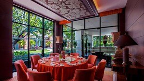 A dining area with a round table set for six, surrounded by orange chairs, large windows offering a view of greenery, and a decorative ceiling.