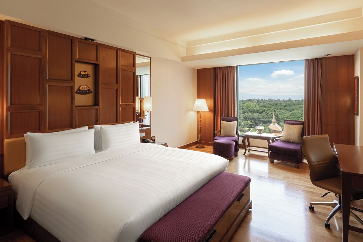 Quarto de hotel com uma cama grande, uma escrivaninha, uma cadeira e vista para uma paisagem arborizada.