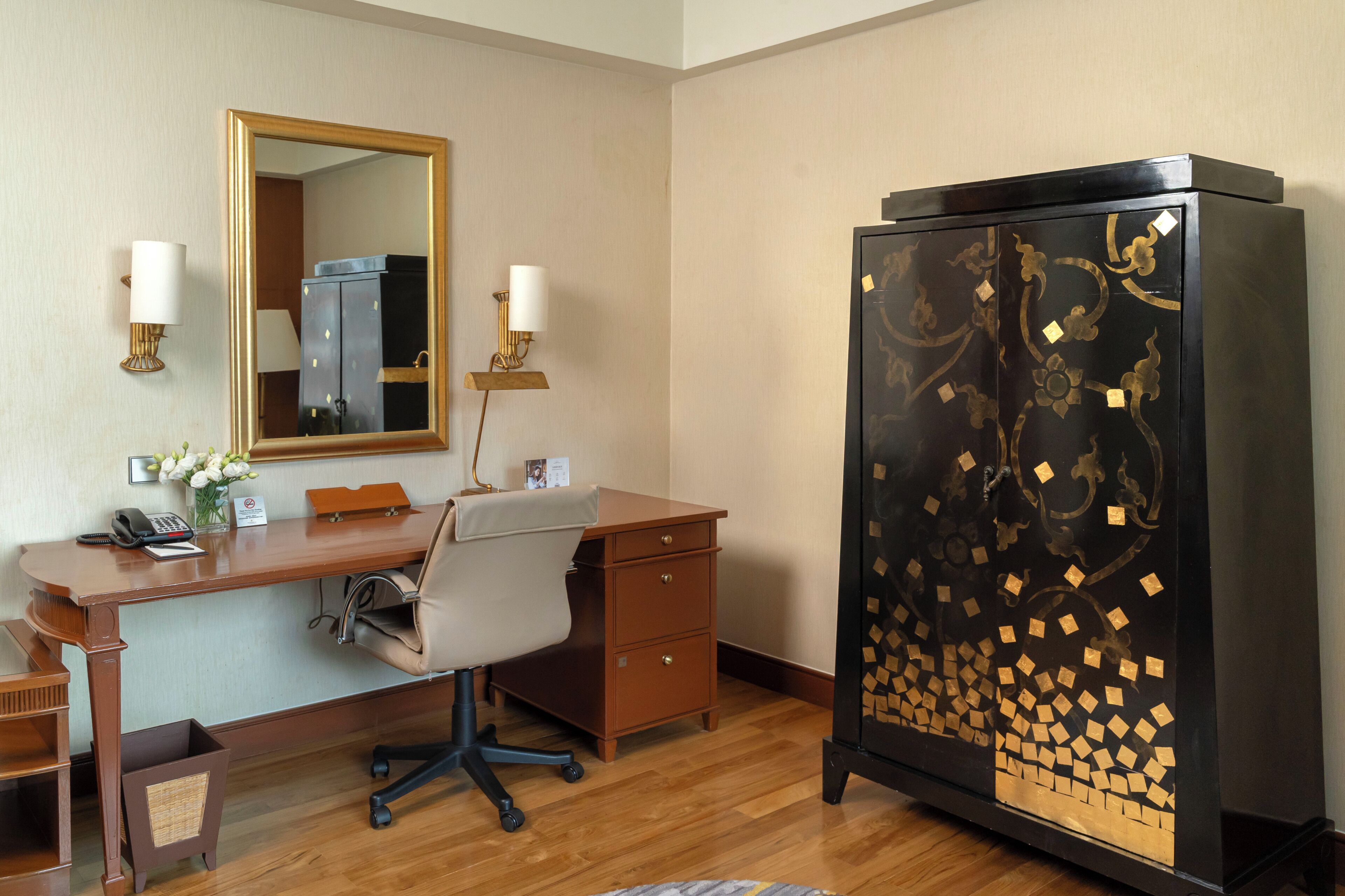 A hotel room with a wooden desk, a chair, a telephone, a mirror, and a cabinet with decorative patterns.