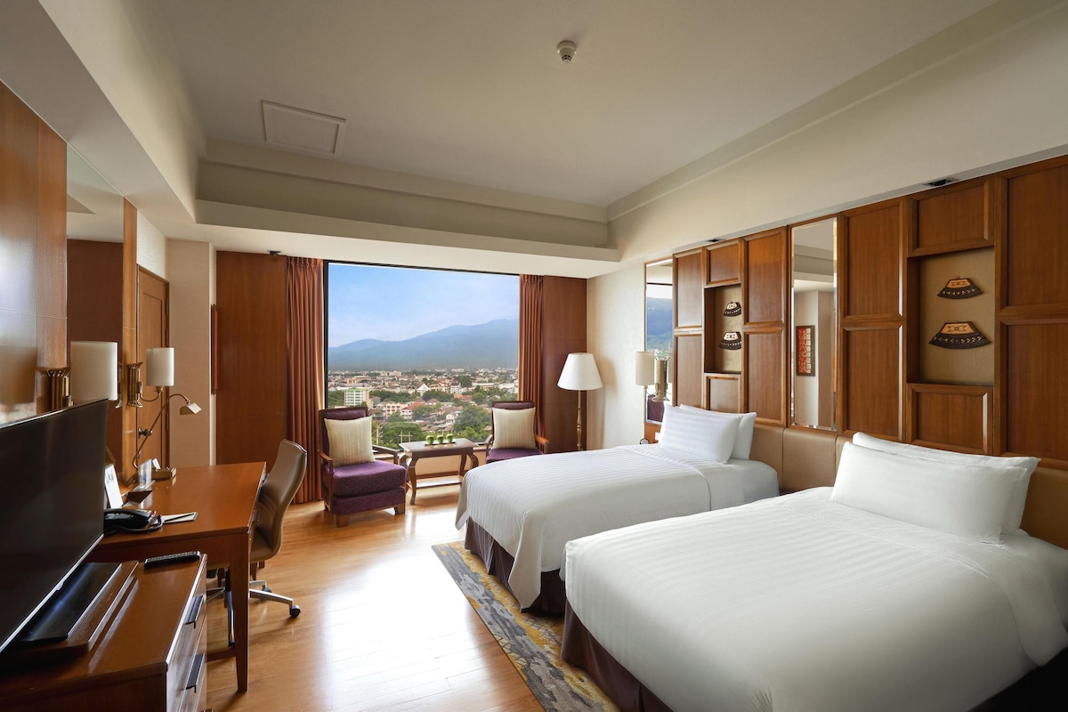 Quarto de hotel com duas camas, uma escrivaninha com televisão, uma cadeira e vista para a cidade, além de painéis de madeira.
