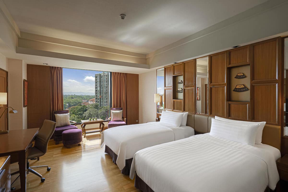 A hotel room with two beds, a desk, a chair, and a view of a cityscape.